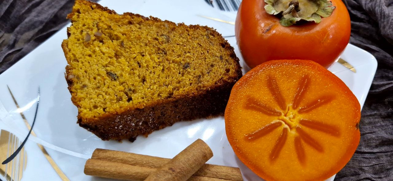 A close-up slice of moist and fluffy persimmon cake on a white plate with fresh persimmon slices, showing the tender and soft crumb