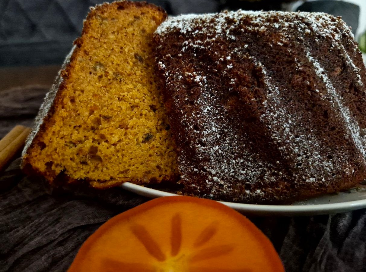 A sliced moist persimmon bundt cake dusted with powdered sugar and a fresh persimmon fruit slice in the foreground